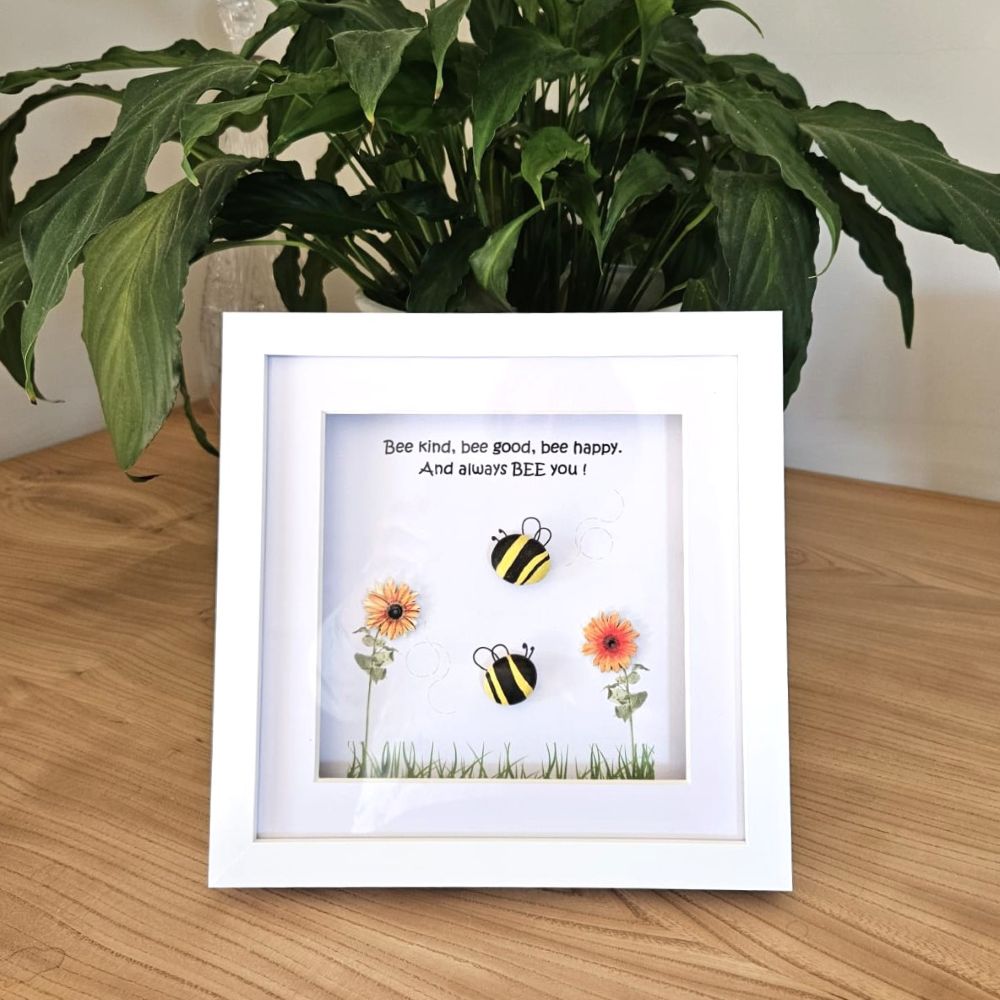 Framed artwork with bees and flowers on a wooden surface with a plant in the background