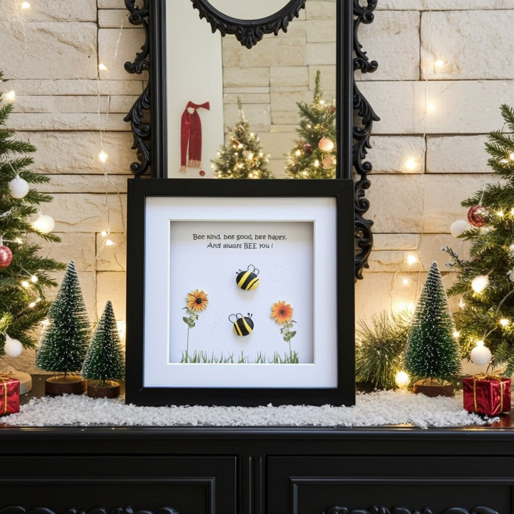 A framed decorative pebble art print displayed on a mantel in a festive holiday setting. The black-framed print features a white background with a be kind quote above two small bumblebees and flowers.
