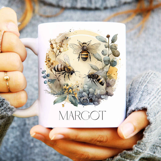 Person holding a mug with bee and floral design, featuring the name 'MARGOT'.