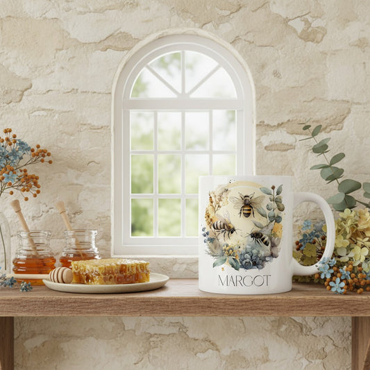 Mug with bee design and 'MARGOT' text on a wooden surface with honey and flowers.