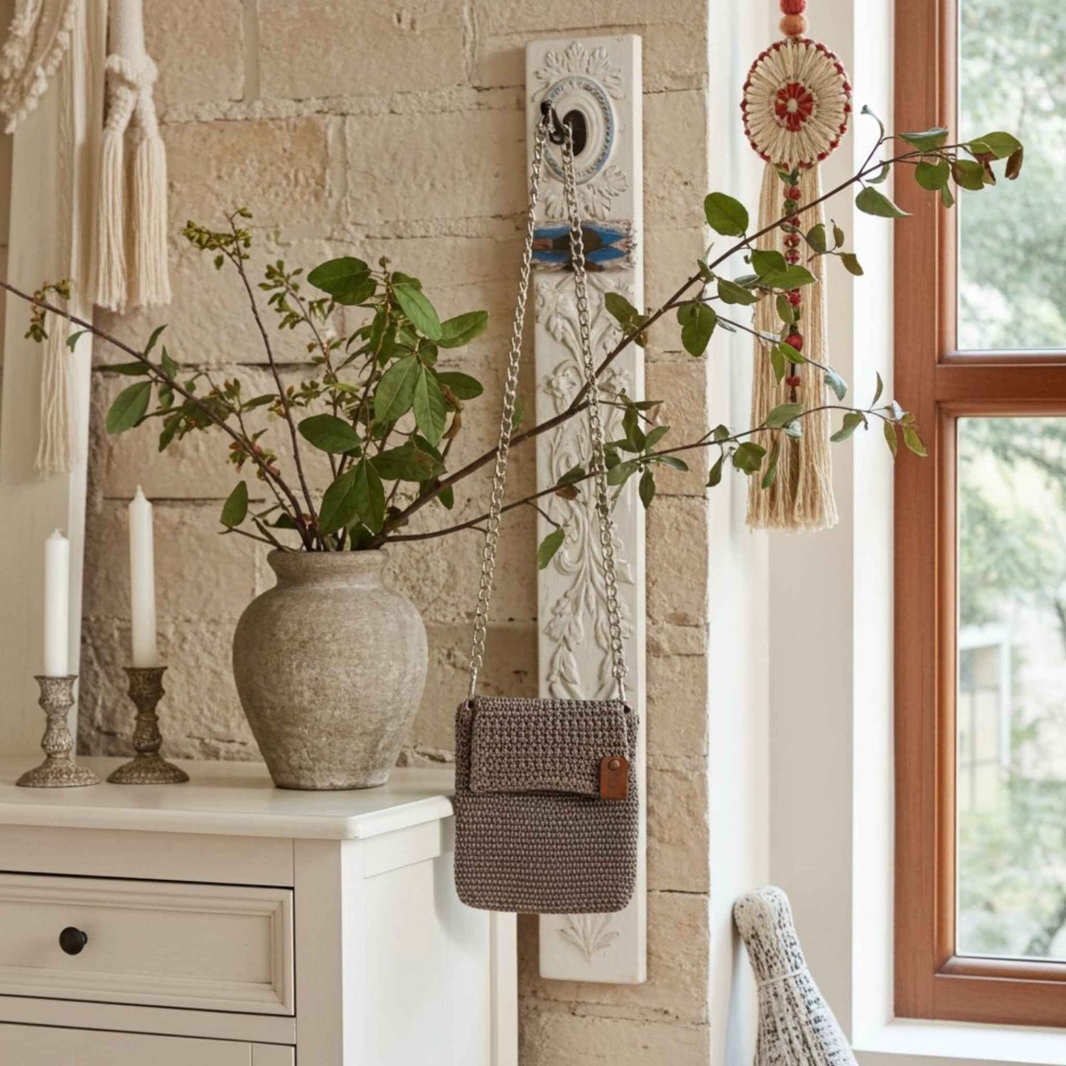 Decorative wall hanging with a vase, branch, and small crochet phone bag against a stone wall.