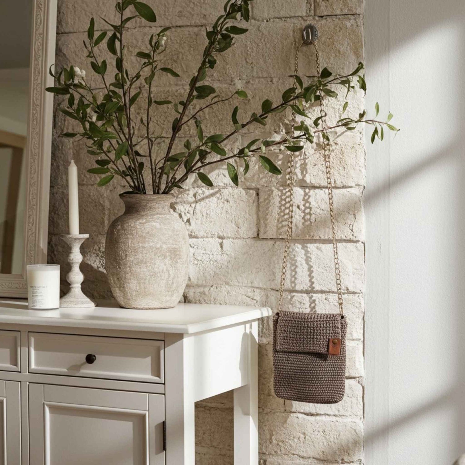 Decorative setting with a vase of greenery, candle, and woven crochet bag against a stone wall.