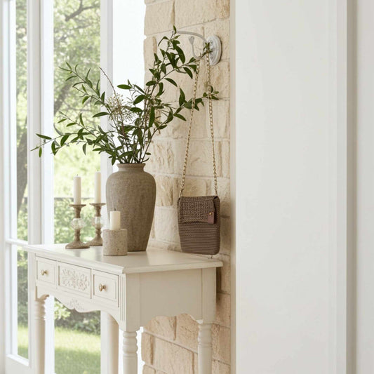 Decorative setting with a vase of greenery, candles, and a beige crochet handbag on a white surface near a window.