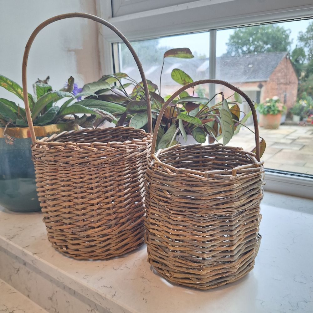 Two handmade willow berry picking baskets with handles, placed on a windowsill, with plants inside them.