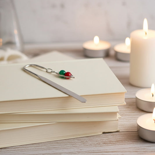 A silver metal bookmark shaped like a planet orbiting two small beads, one green and one red, resting on a stack of cream-coloured books. The scene is styled with warm, glowing tea light candles on a light wooden surface.
