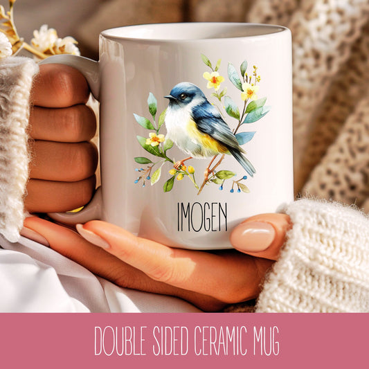 A double-sided ceramic mug with a bird design and the name 'Imogen' printed on the other side, held in a hand with a floral knit hat and a cozy grey sweater in the background.