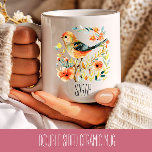 A double-sided ceramic coffee mug with an illustration of a bird and flowers, and the name Sarah printed on it.
