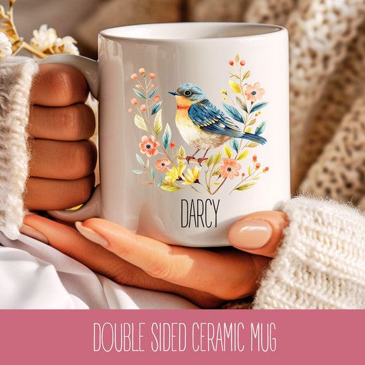 A hand holding a double-sided ceramic mug with a bird and floral design, with the name 'Darcy' printed on it.