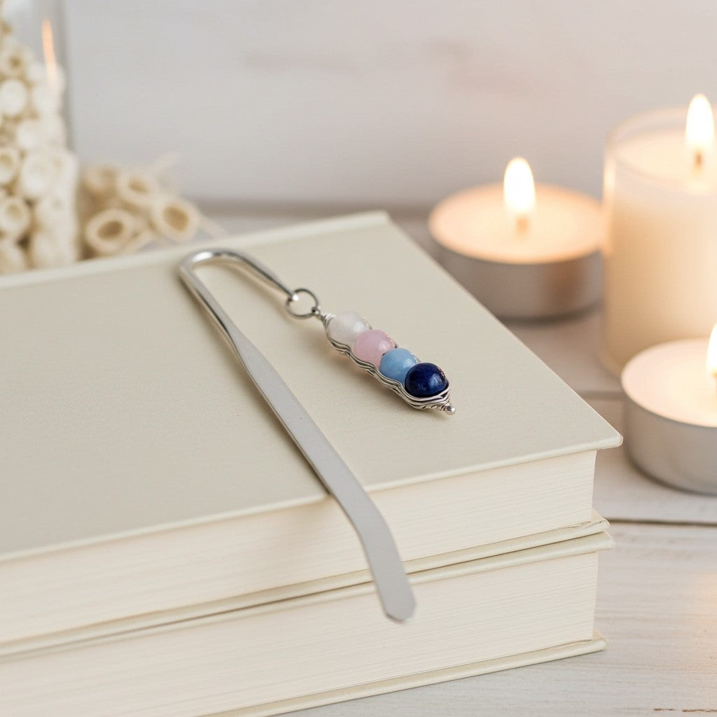 A silver metal bookmark with three small coloured beads (white, pink, and dark blue) resting on a stack of cream-coloured books, with warm, glowing candles blurred in the background.