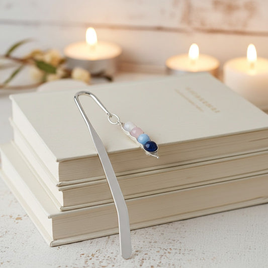 A silver metal bookmark with three small beads in pastel pink, light blue, and dark navy, resting on a stack of cream-coloured books with blurred lit candles in the background.