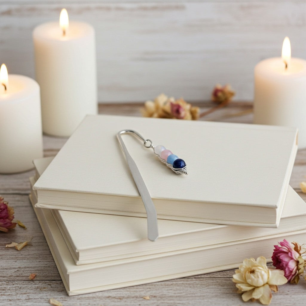 A silver metal bookmark with pastel pink, light blue, and dark blue beads rests on a stack of cream-coloured hardcover books, styled with lit white candles and dried flowers on a rustic wooden surface.