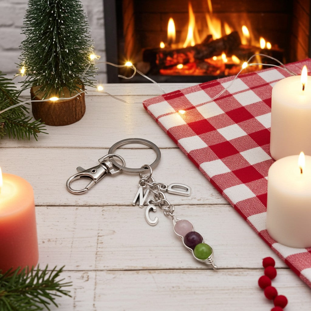 A silver keychain with a lobster clasp, a keyring, and three hanging charms including a silver 'M' initial, a 'C' initial, and a beaded charm with pink, purple, and green beads, styled on a white wooden table with a red and white checkered cloth, candles, a small Christmas tree, and a fireplace in the background.