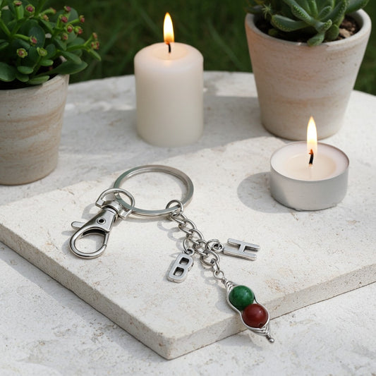 A silver-toned metal keychain with a lobster clasp, key ring, and small hanging charms, including a green and red beaded charm, displayed on a light stone surface with lit candles and potted plants in the background.