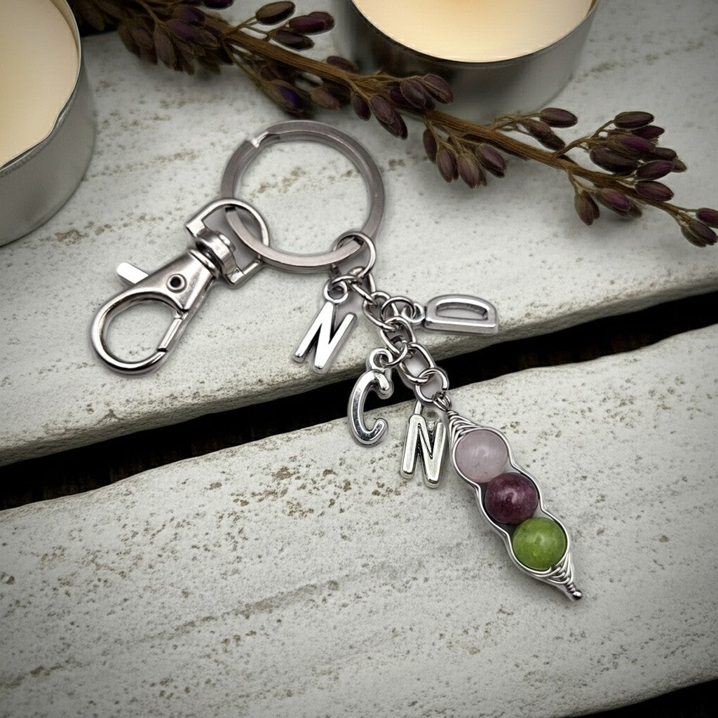 A silver-toned keychain with a lobster clasp and dangling letter charms, including 'N', 'C', and 'N', plus a small bead-and-branch charm in pink, purple, and green, photographed on a light stone surface with candles and dried stems in the background.