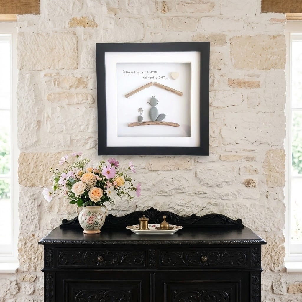A framed piece of wall art featuring a pebble cat and wooden house design  displayed on a rustic stone wall above a dark wooden console table with a floral arrangement.