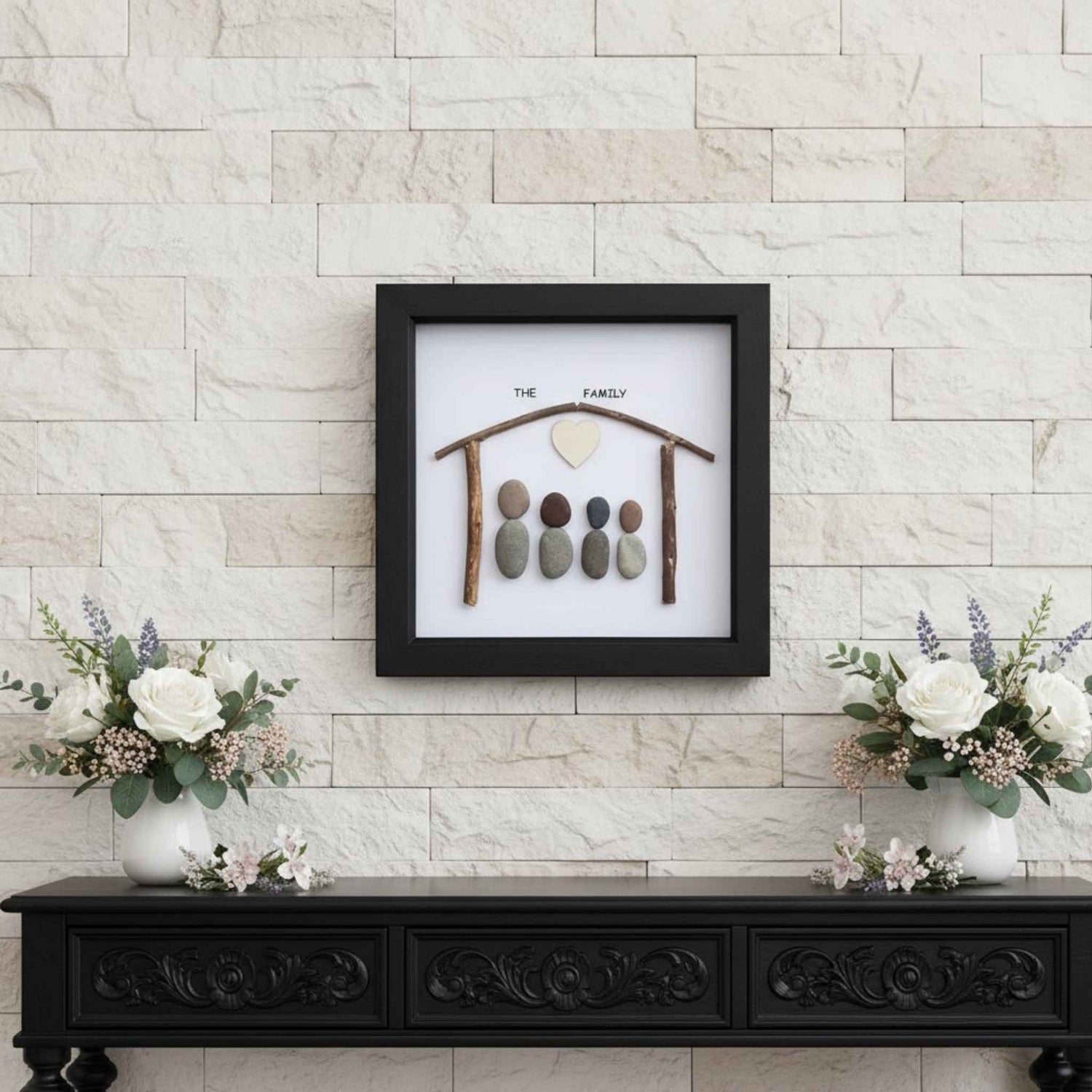 Decorative wall art with pebbles and branches on a white background, framed in black against a stone wall.
