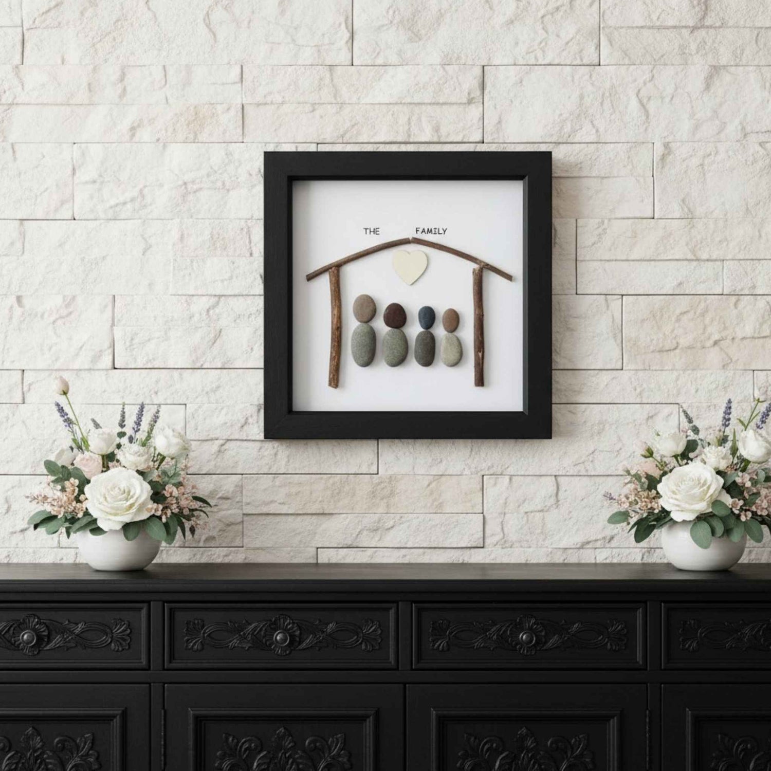Framed artwork with pebble family design on a wall above a black cabinet with floral arrangements.