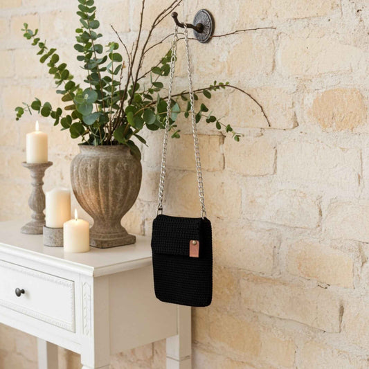 Black woven phone handbag hanging on a wall hook next to a white cabinet with candles and a plant.