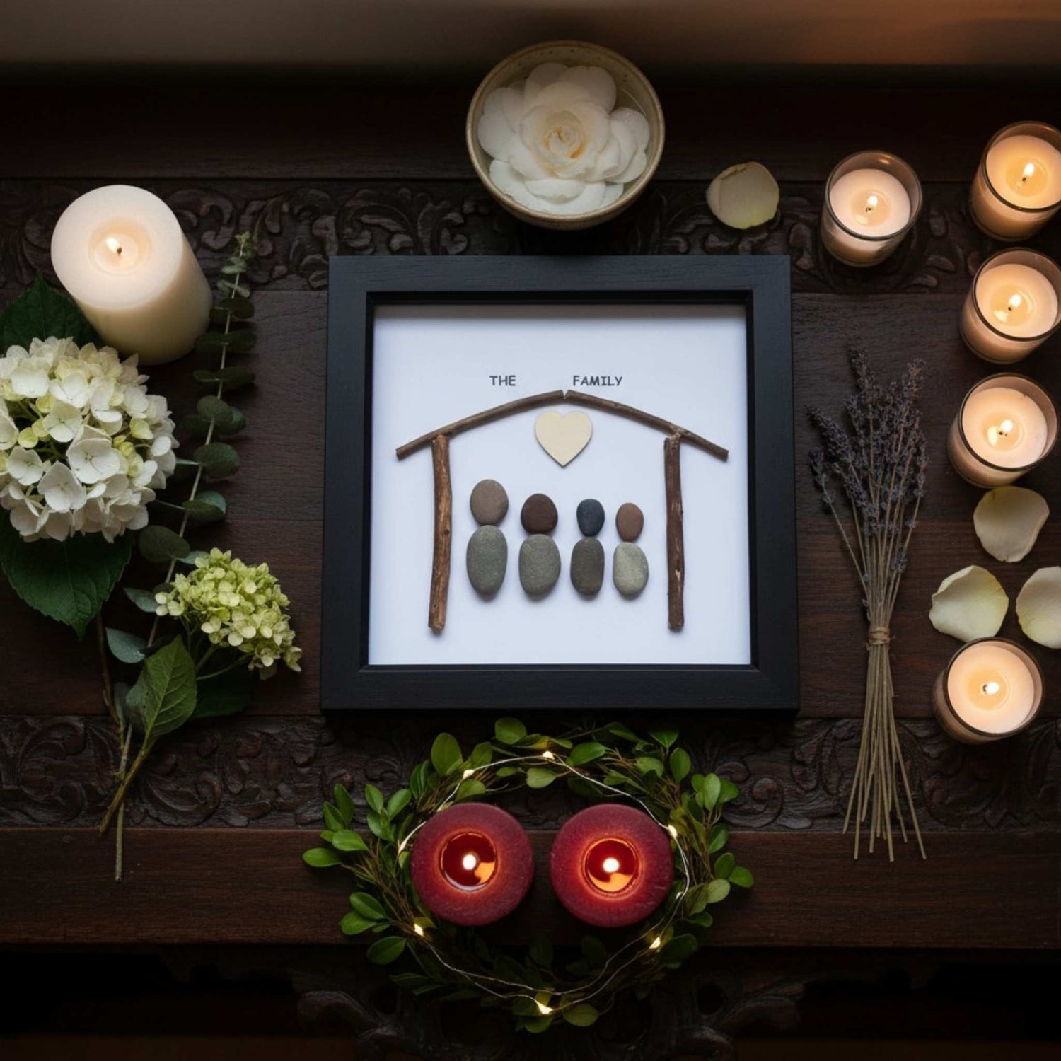 Decorative pebble family portrait in a black frame surrounded by candles and flowers on a wooden surface.