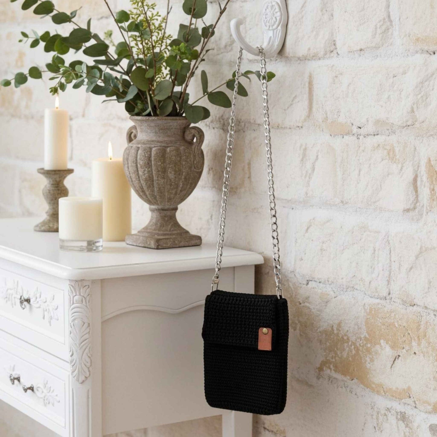 Black woven phone handbag hanging on a hook next to a white dresser with candles and an urn.