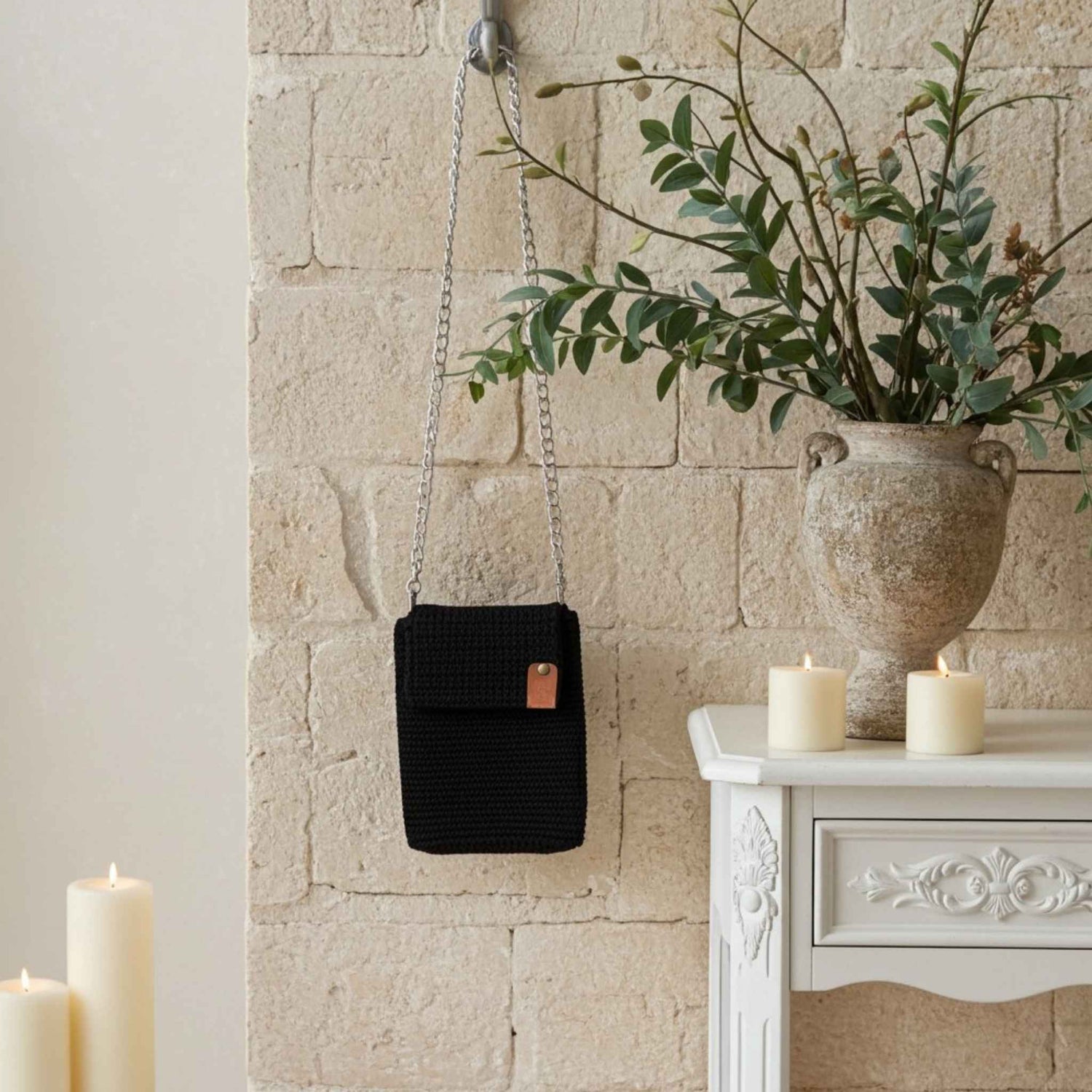 Black woven phone handbag with a chain strap hanging on a stone wall next to a vase with greenery and candles.