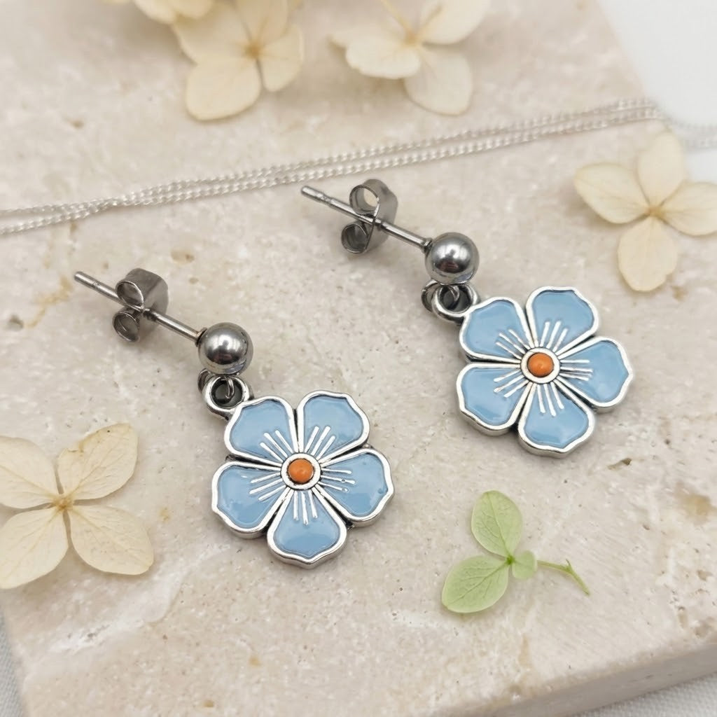A pair of silver-tone dangle earrings with light blue flower charms and small orange centres, displayed on a beige stone surface with dried flowers and a silver chain.