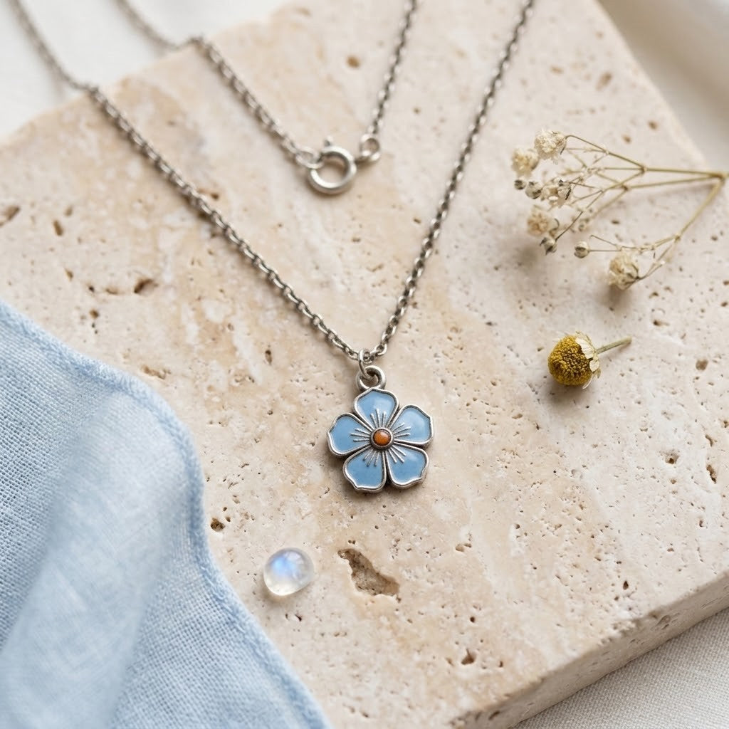 A silver-tone chain necklace with a light blue flower pendant featuring a small gold centre, styled on a beige stone slab with dried flowers and a blue fabric accent.