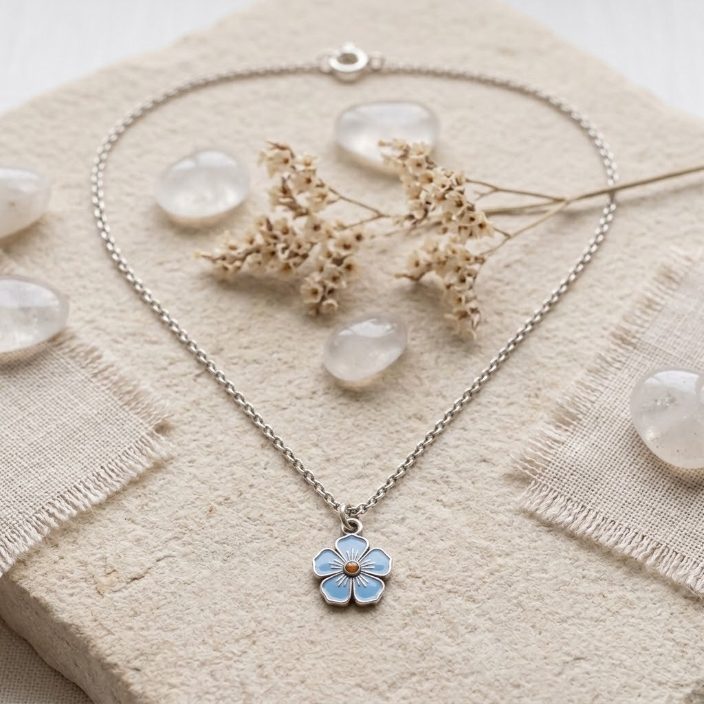 A silver-tone chain necklace with a small flower-shaped pendant featuring light blue enamel petals and a tiny gold centre, styled on a textured beige fabric with dried flowers and clear stones.