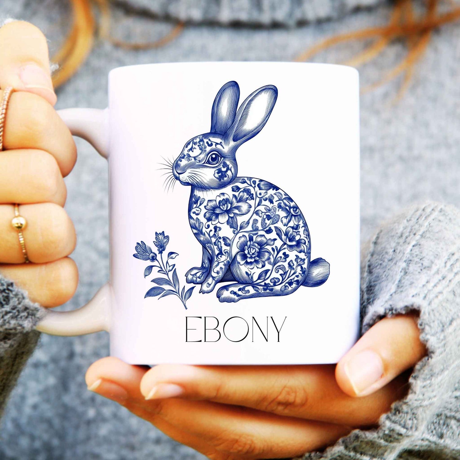 White mug with hare rabbit design held by a person wearing a grey sweater.