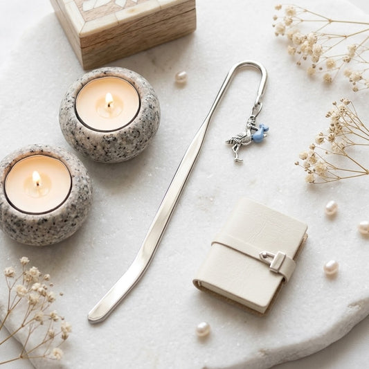 A silver metal bookmark with a small blue stork charm and white pearl accents, styled on a white stone surface with two lit candles, dried flowers, pearls, and a small cream notebook.