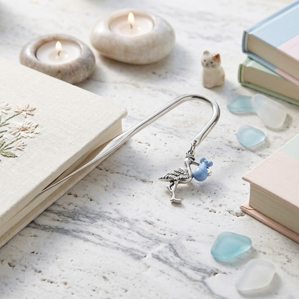 A silver metal bookmark with a blue stork charm, resting on a cream-coloured book on a marble-like surface with candles, stones, and pastel books around it.