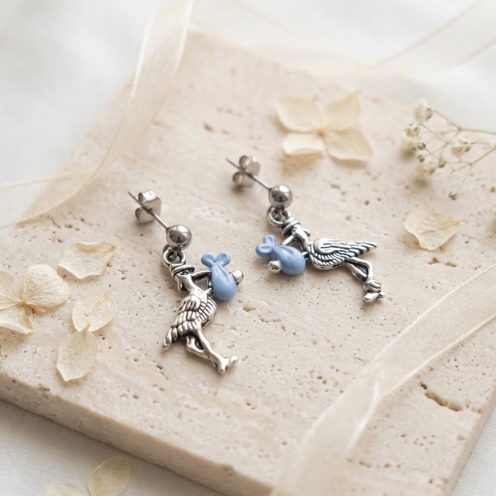 A pair of silver dangle earrings with bird charms holding small light blue pouches, displayed on a beige stone slab with dried flower petals and a sheer ribbon.