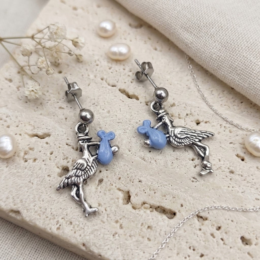 A pair of silver-toned stork earrings with light blue enameled pouch charms, displayed on a textured beige stone slab with decorative pearls and dried flowers.