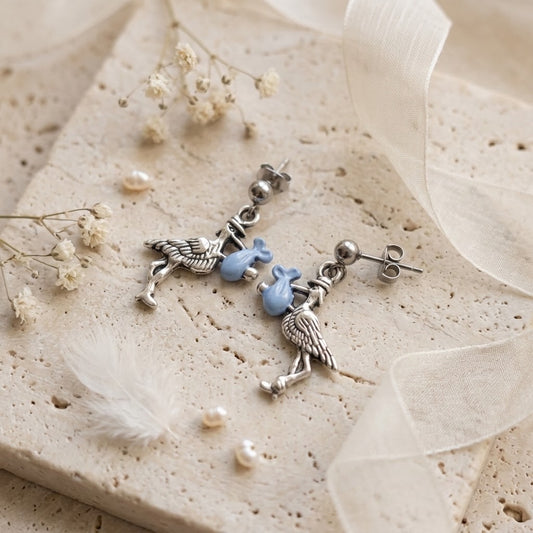 A pair of silver-toned dangle earrings with bird charms and light blue bead accents, styled on a textured beige stone slab with dried flowers, pearls, and sheer white ribbon.