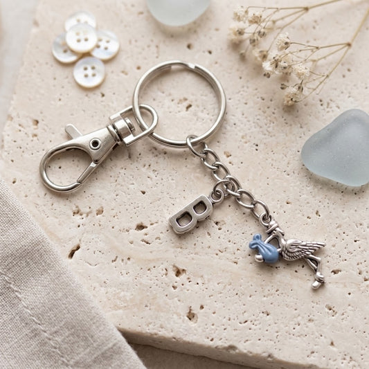 A silver-toned metal keychain with a lobster clasp, split ring, a letter B charm, a bird charm, and a small blue accent, styled on a textured beige stone surface with decorative buttons, dried flowers, and sea glass.