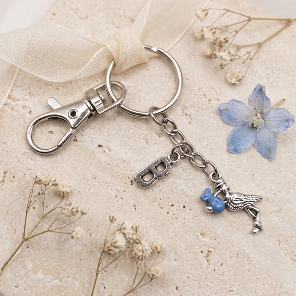 A silver-toned metal keychain with a lobster clasp, split ring, engraved letter charm, and a dangling bird charm holding a small blue bead, styled on a light beige textured surface with dried flowers and a sheer cream ribbon.