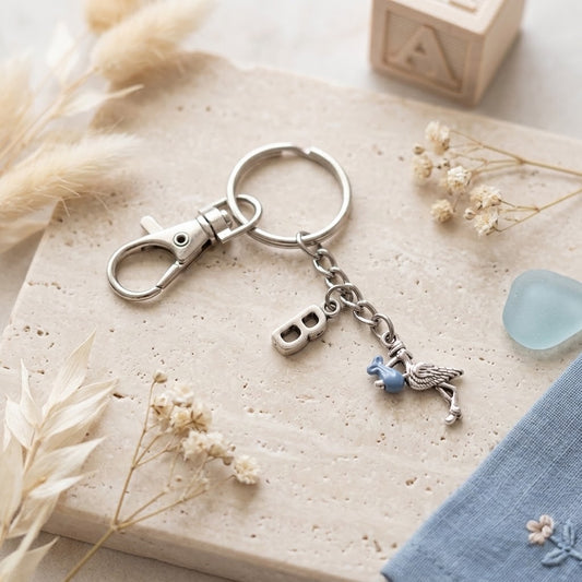 A silver-toned metal keychain with a lobster clasp, split ring, letter B charm, and a small blue bird charm, styled on a textured beige stone slab with dried flowers and a blue fabric corner.