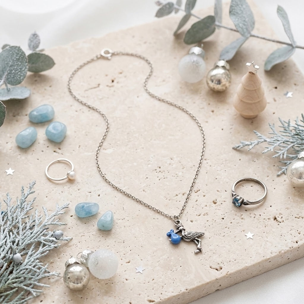 Silver-toned necklace with a small blue bird charm, styled on a beige stone slab with rings, blue stones, silver ornaments, and eucalyptus branches.