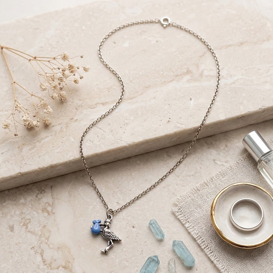A silver-toned chain necklace with a blue stork pendant holding a small blue flower, styled on a light beige stone slab with dried flowers, crystals, and a perfume bottle nearby.