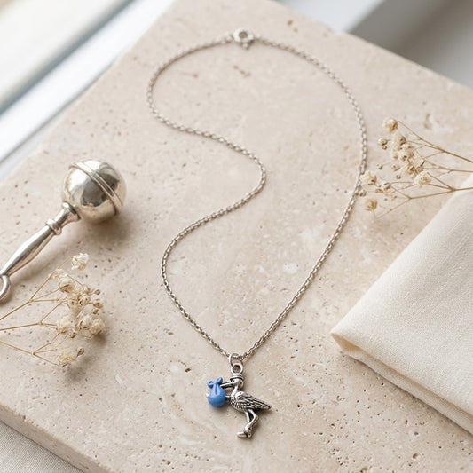 A silver-toned chain necklace with a stork charm holding a small blue flower, styled on a beige stone slab with dried flowers and a folded cream fabric.