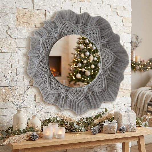 A round decorative wall mirror with a large scalloped grey macrame border hangs on a light stone wall above a wooden console table decorated with candles, pinecones, wrapped gifts, and greenery. The mirror reflects a lit Christmas tree in a cozy indoor setting.