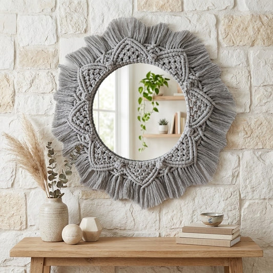 A round wall mirror with a large grey macrame-style fringe border, mounted on a light stone wall above a wooden console table with decorative vases, books, and a bowl.