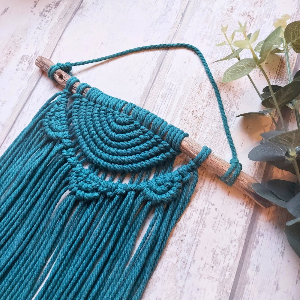 A turquoise boho macrame wall hanging with a semi lunar moon phase design, displayed on a white wooden background with green foliage in the top right corner.