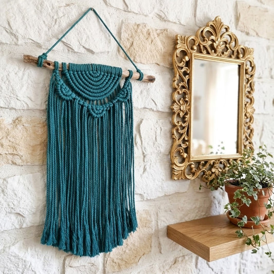 A teal macrame wall hanging suspended from a wooden dowel, displayed on a white stone wall next to an ornate gold mirror and a small wooden shelf with a potted plant.