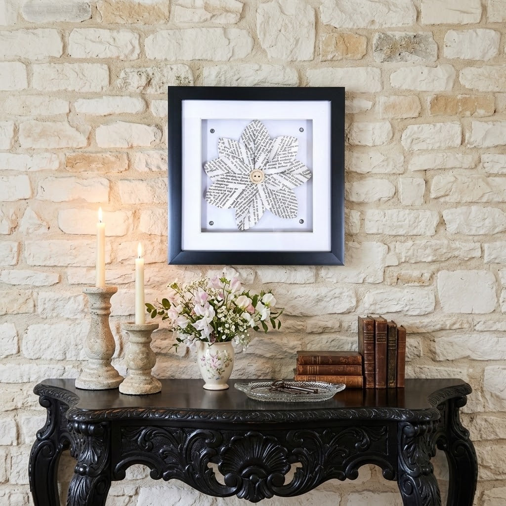 A square framed wall art piece featuring a book page flower design on a white background, hung on a light stone wall above a dark carved console table with candles, flowers, and books.
