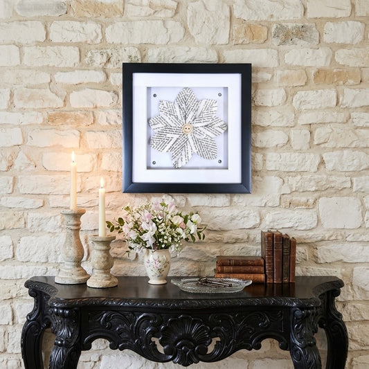 A square framed wall art piece featuring a book page flower design on a white background, hung on a light stone wall above a dark carved console table with candles, flowers, and books.
