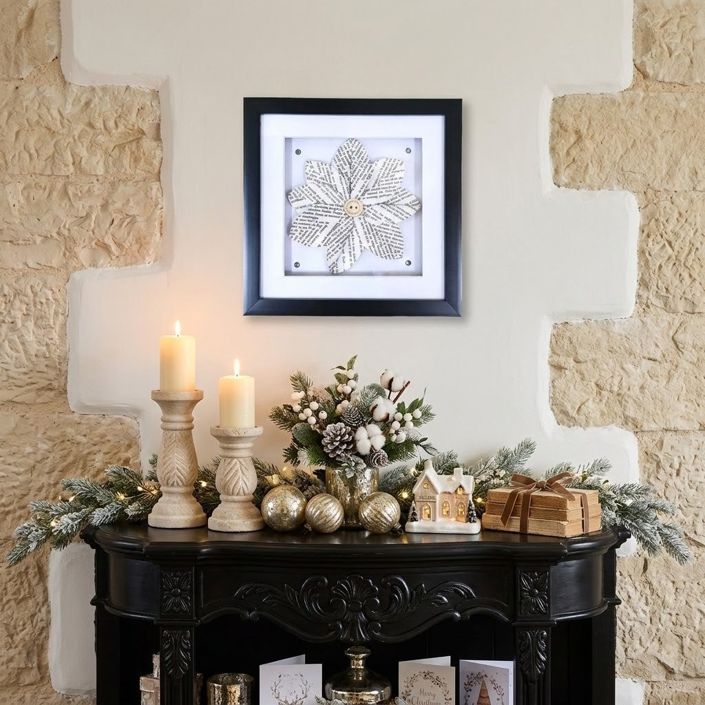 A framed square wall art piece featuring a floral page from a book, displayed on a beige wall above a dark wooden console table decorated with candles, pinecones, and holiday ornaments.