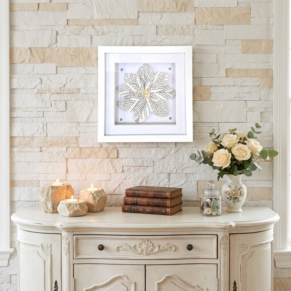 A white square shadow box frame displaying a book page flower design, mounted on a light beige stone wall above a distressed cream console table decorated with candles, books, and a vase of roses.