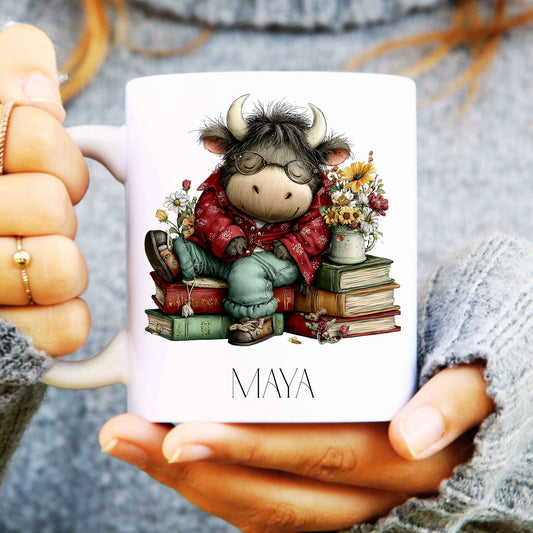 Person holding a mug with a cartoon highland cow character and 'MAYA' text.