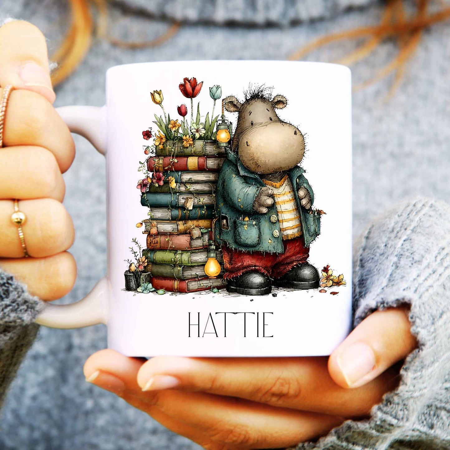 Person holding a mug with a cartoon hippo and books design, labeled 'HATTIE'.